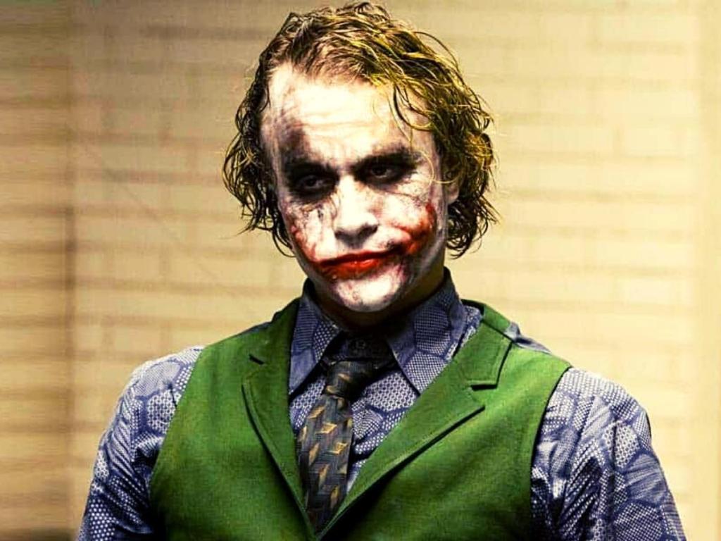 Heath Ledge in "The Dark Knight" in full makeup and costume as the Joker. He has greasy green-tinged blondish hair, white clown face paint, black grease paint around his eyes, and red grease paint on his lips and up his cheeks to make a smile. He's wearing a purple hexagon-print shirt and a green waistcoat.