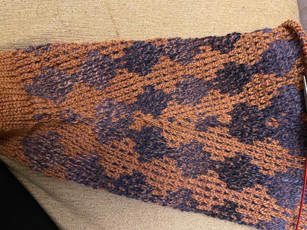 A photo of an unfinished knitted sweater sleeve made with multicolor purple yarn and brown yarn. The pattern is a diamond motif, and as the sleeve increases in size, more diamonds are added and appear to "burst" from the origin point where the increases occur in a symmetrical pattern.