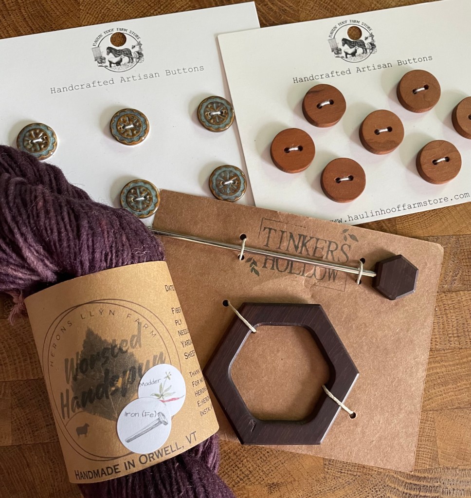 Top of the photo shows two white post cards with circular buttons fastened to them - one set is turquoise with a light brown floral design, and the other set is light brown wood. In the bottom left corner is a hank of dark purple yarn with a brown paper label; on the right is a piece of brown cardstock with a dark brown wooden hexagon fixed to it, with a matching pin.