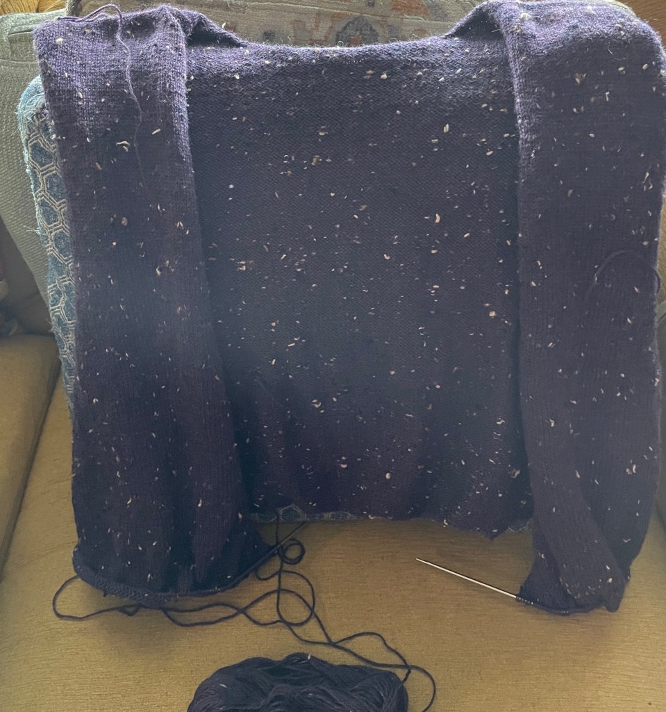 The front of unfinished knitting project of a cardigan made with dark purple yarn with tweedy neps. The project is draped over a stack of throw pillows on a yellow couch, and the string of yarn connects to mostly-used skein at the bottom center of the frame.