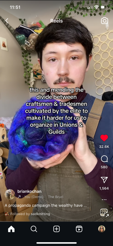 A screenshot of Brian's video. He holds up an unspun batt of royal blue fiber. He looks directly in the camera, and over the image is text that reads "This and mending the divide between craftsmen & tradesmen cultivated by the elite to make it harder for us to organize in Unions & Guilds". Brian has light skin, dark brown hair and eyes, and a mustache. He wears a blue shirt and brown overalls.
