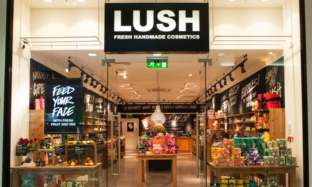 A storefront of Lush, which sells skin and hair care products as well as perfumes and bath bombs. The signage is black with white text, the interior is made entirely from wood, and the displays are filled with colorfully wrapped soaps and boxes.
