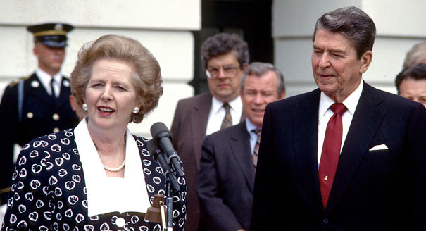 Margaret Thatcher stands on the left wearing a dark blue printed garment with a wide white collar. She's wearing a pearl necklace and large pearl earrings and is standing in front of a microphone. Ronald Reagan stands to her right, wearing a dark blue suit and a red tie.