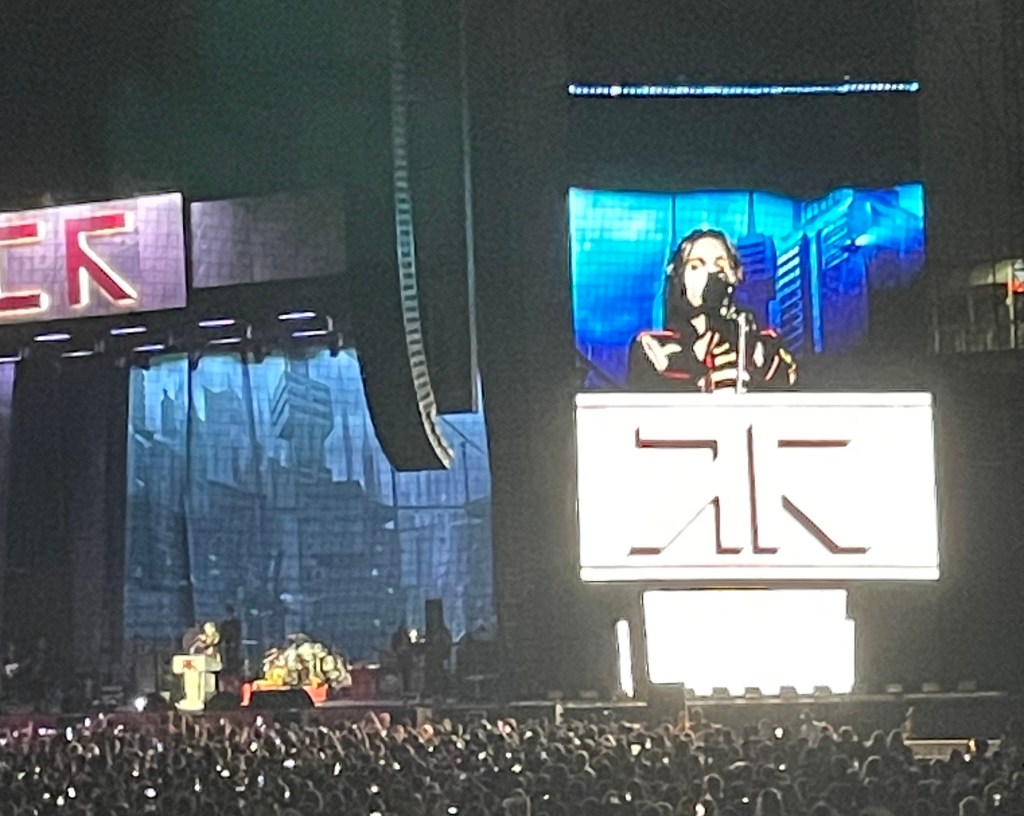 On the left side, the band plays on the stage; on the right, GerardWay appears on the jumbotron behind a podium.
