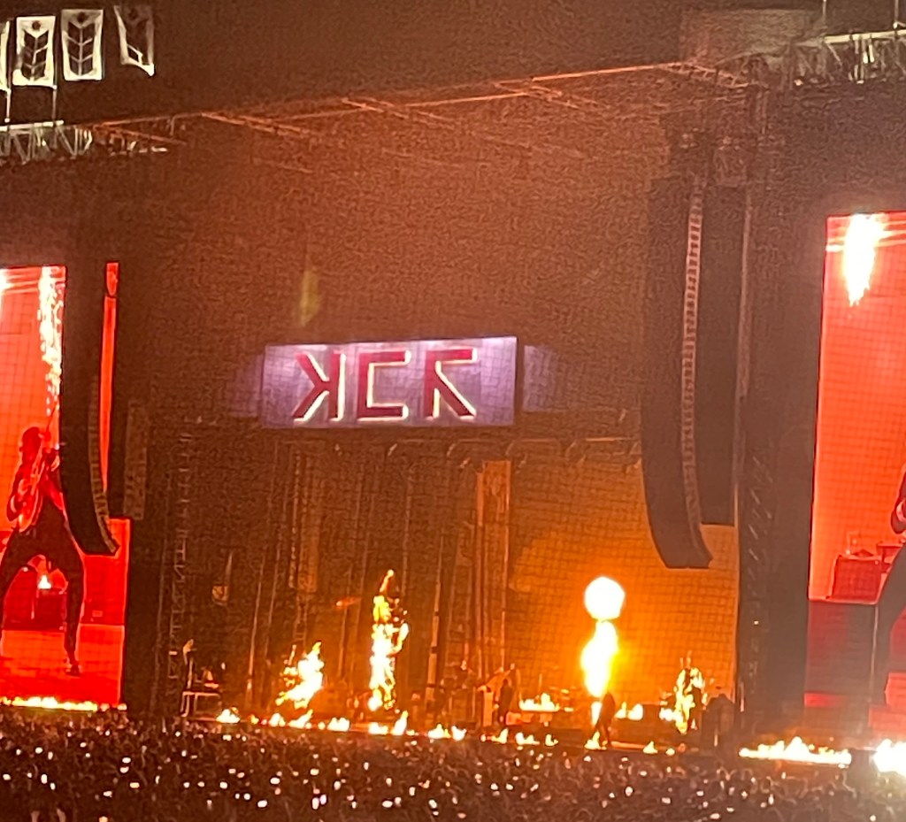 A wide shot of the stage. There is fire at the edge of the stage, in the back, and the banners on either side of the band are catching fire. Everything is orange, black, and red.