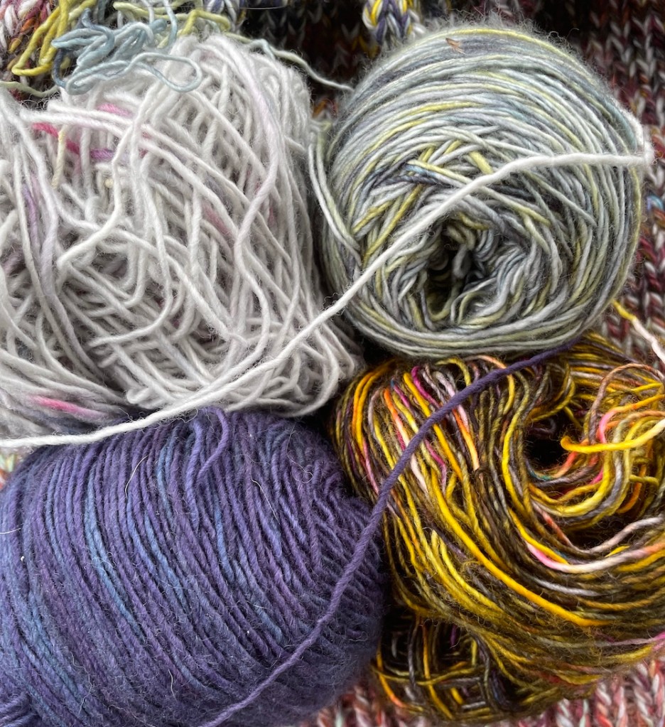 Four balls of yarn. From clockwise bottom left: A deep blue and purple yarn, a silver yarn with pink speckles, a light green yarn speckled with yellow and dark blue gray, and a multicolor yarn with brigh yellow, hot pink, and blank.