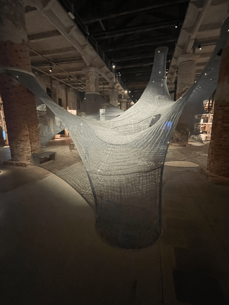A large knitted art piece is suspended on multiple brick columns in a large room, kind of like a giant spiderweb. Multiple lines of LED lights run through it, and dots of light can be seen throughout.