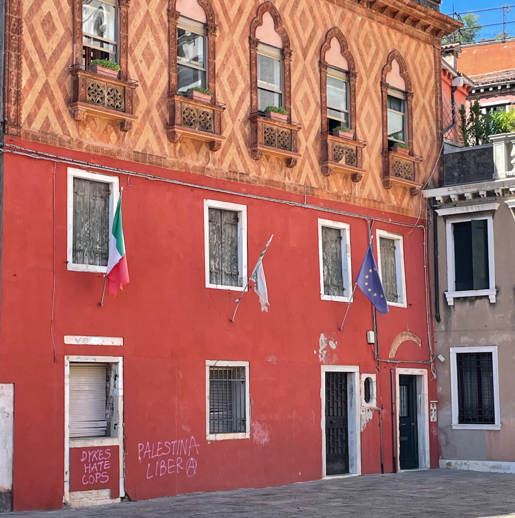 A building whose first floor is painted orangey-red and whose top floor has an intricate, diamond brick design. In the lower left corner the words "Dyles hate cops" and "Palestina libera" have been spray-painted in pink.