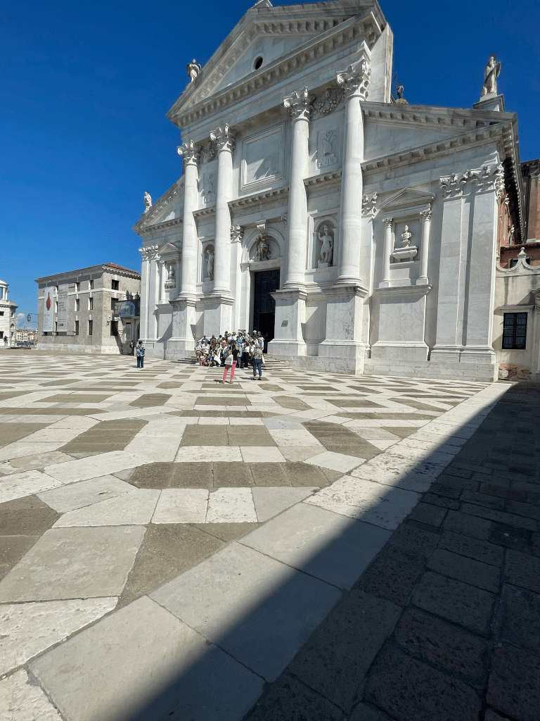 The facade of San Giorgio Basilica, a white marble building with a stone plaza, tiled in a geometric pattern. 