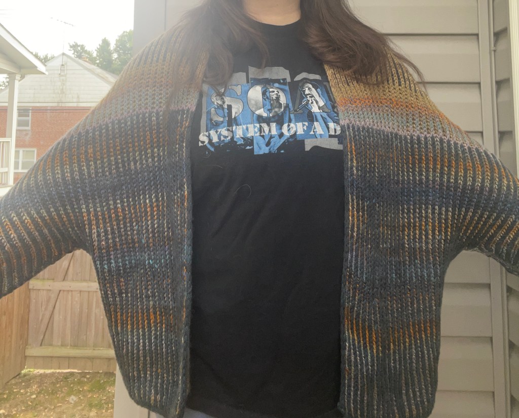 I'm standing in front of my house's wall wearing the Oliveros, which I've knit in antique gold, navy, and a multi-color yarn with blue and orange in it. I'm wearing a black System of a Down t-shirt with blue and white text. The photo is cropped to only show my shoulders down to my hips.