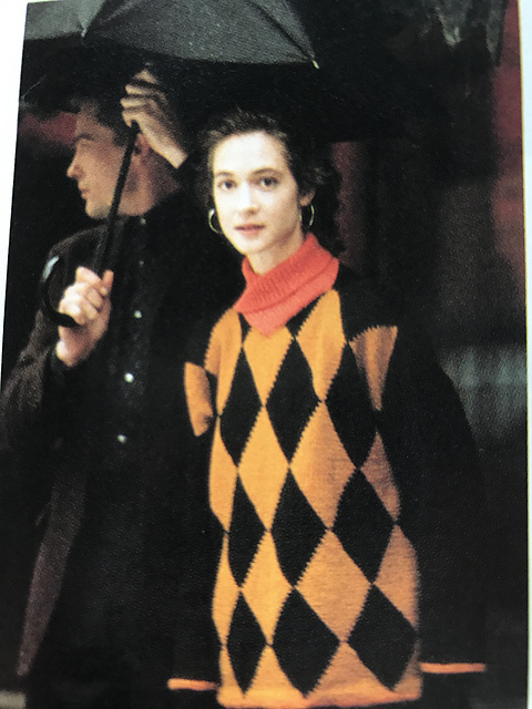 A brunette model stands wearing the Harlequin Pullover, which has light orange and black diamons on the torso, black sleeves, and a coral pink cowl collar. She stands in front of a male model holding an umbrella.