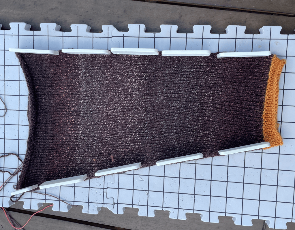 A sleeve, pinned to a white blocking mat. It's in the dark, marled yarn with black, red, gray, orange, and pink. It also has a roll cuff knit in orange yarn.