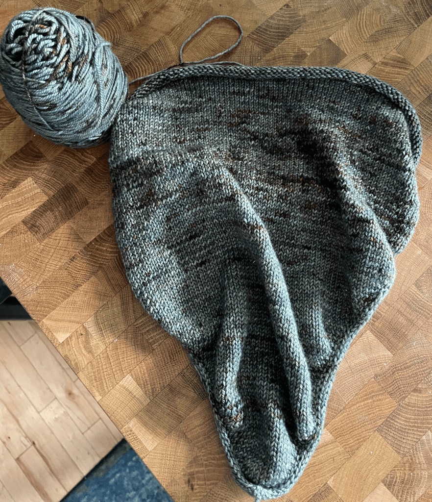 A triangule-shaped, unfinished bandana cowl knit in the green-gray color with rusty brown speckles.