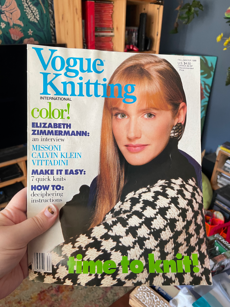 I'm holding up an issue of Vogue Knitting magazine. My hand is visible in the lower left of the photo. The magazine cover features a blonde model with bangs wearing a black and white sweater that's knit in a houndstooth pattern. The title of the magazine is in turquoise blue, and at the bottom it reads "Time to knit!" in lime green. The background of the cover is bright white.